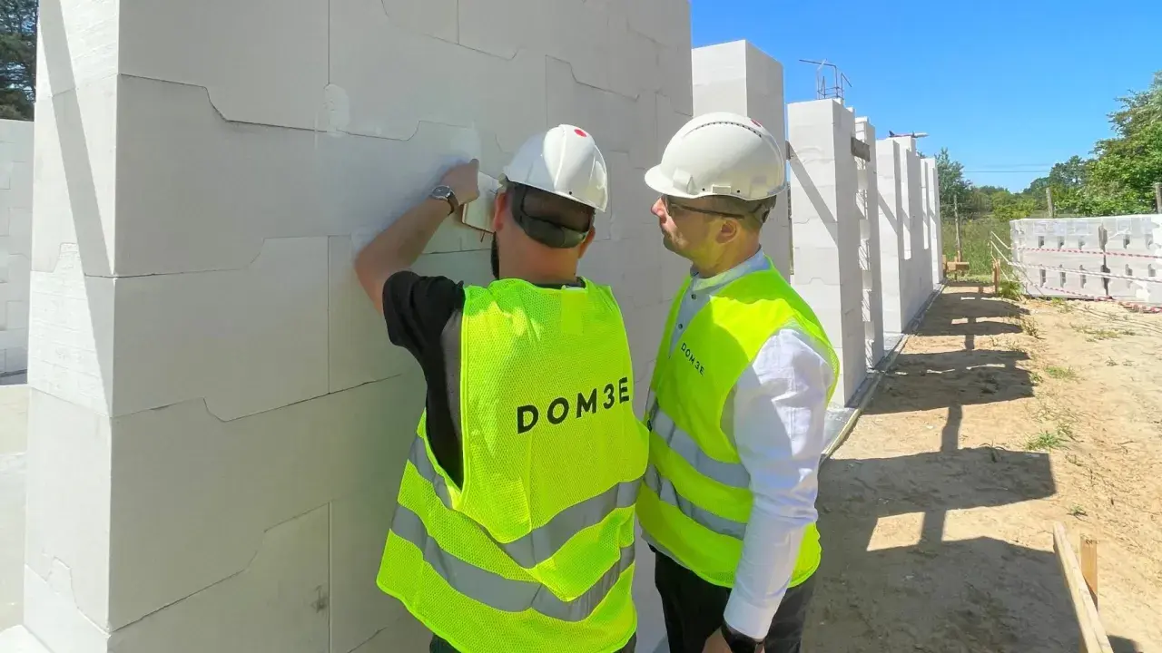 Dwóch mężczyzn w kamizelkach odblaskowych i kaskach podczas inspekcji budowlanej. Jeden z nich sprawdza bloczki z betonu komórkowego.