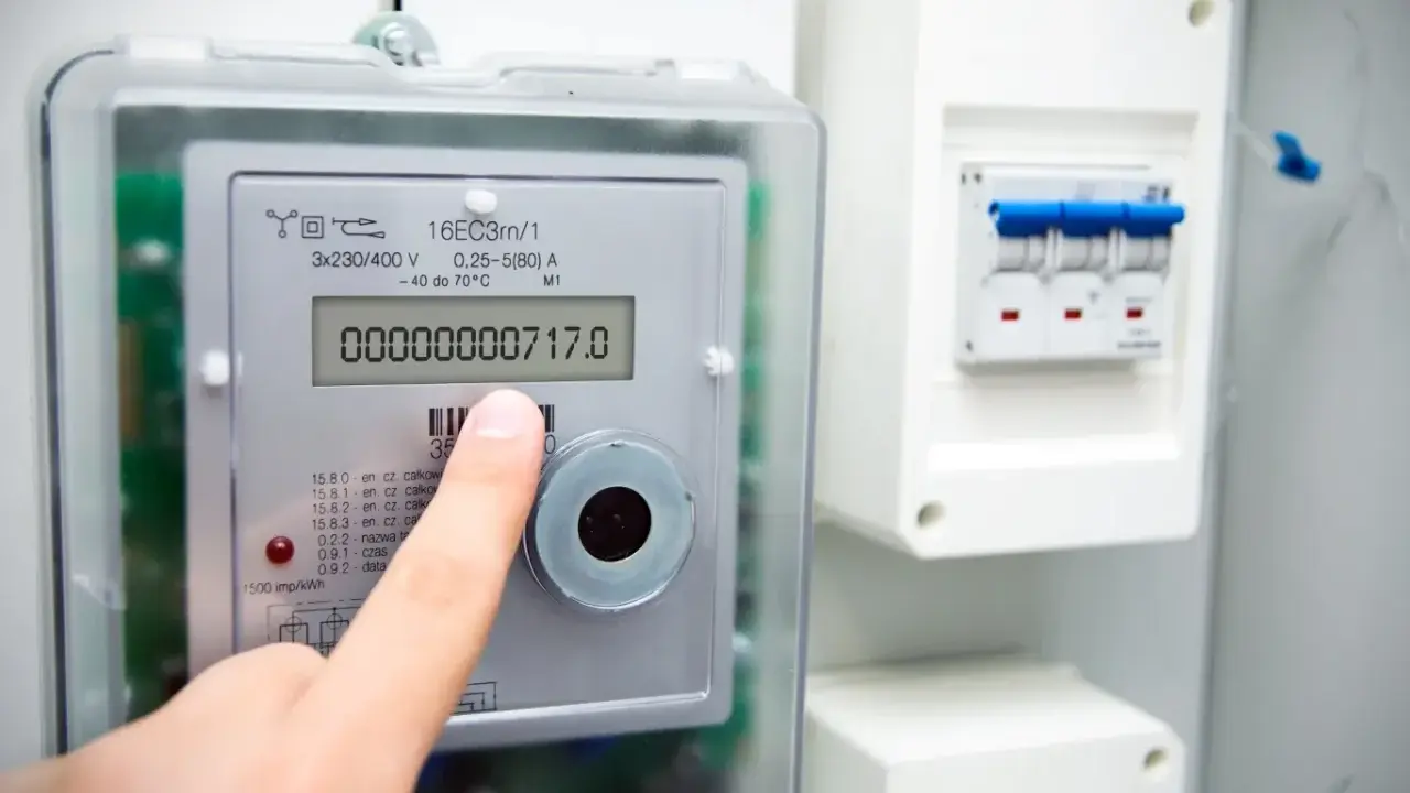 Palec wskazuje stan licznika energii elektrycznej: 00000000717.0 kWh. Obok widoczny wyłącznik.