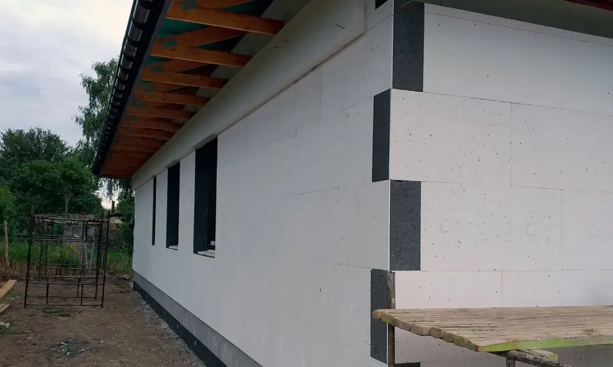 Budowa domu w toku. Ściany ocieplone grubym styropianem na placki, widoczne drewniane elementy dachu i okna.