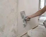 Ręka z kielnią nakłada gładź na chropowatą ścianę, pokazując, jak położyć gładź na ścianę dla idealnie równej powierzchni.