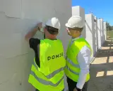 Dwóch mężczyzn w kamizelkach odblaskowych i kaskach podczas inspekcji budowlanej. Jeden z nich sprawdza bloczki z betonu komórkowego.