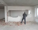 Mężczyzna wyburza ścianę działową, tworząc otwór. Widać gruz i cegły.