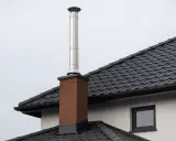 Nowy komin w starym domu, pozwolenie na budowę zostało wydane. Stalowa rura na ceglanej podstawie na tle szarego nieba.