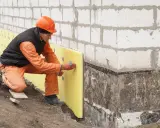 Budowlaniec mocuje żółty styropian do ściany fundamentowej. Czy styropian pije wodę? Nie, jest wodoodporny.