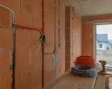 Instalacja elektryczna w trakcie budowy. Widać pomarańczowe peszle i kable prowadzone po ceglanej ścianie. Na pierwszym planie zwoje kabli i wkrętarka.