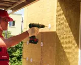 Pracownik w kasku montuje wełnę mineralną do ściany drewnianej za pomocą wkrętarki.