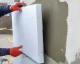 Ręce w czerwonych rękawicach przytwierdzają biały styropian do ściany. To dowód na świetne właściwości styropianu w izolacji termicznej.