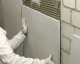 Ocieplanie ściany od wewnątrz: pracownik w białej kurtce i rękawicach przykłada płytę izolacyjną do ściany z zaprawą.