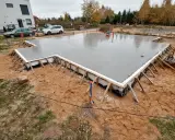 Świeżo wylana ława fundamentowa na budowie domu. Wokół plac budowy z piaskiem i trawą.