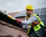 Instalator w żółtym kasku i kamizelce odblaskowej montuje panele fotowoltaiczne na dachu. Czy fotowoltaika jest bezpieczna? Tak, gdy montaż wykonują profesjonaliści.