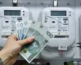 Dłoń trzyma polskie banknoty przed licznikiem energii elektrycznej. Brak licznika dwukierunkowego przy instalacji fotowoltaicznej może oznaczać brak rozliczenia nadwyżek.