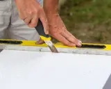 Ręce trzymają nóż i poziomicę, pokazując, jak przeciąć styropian domowym sposobem.