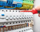Ręka z czerwonym śrubokrętem dokręca śrubę w budowie rozdzielni elektrycznej. Widać bezpieczniki i kolorowe przewody.