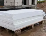 Stos białych płyt styropianowych na palecie, stojący na mokrej ziemi. Czy styropian może leżeć na deszczu? Materiał budowlany na placu budowy.