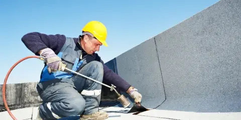 Pracownik w żółtym kasku wykonuje zgrzewanie papy na dachu, używając palnika.