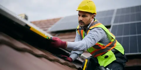 Instalator w żółtym kasku i kamizelce odblaskowej montuje panele fotowoltaiczne na dachu. Czy fotowoltaika jest bezpieczna? Tak, gdy montaż wykonują profesjonaliści.