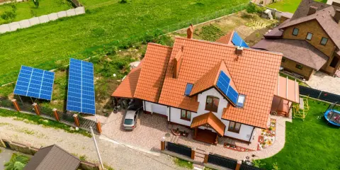 Dwa panele fotowoltaiczne na ziemi i kilka na dachu domu. W tle zielone pole i sąsiednie budynki.