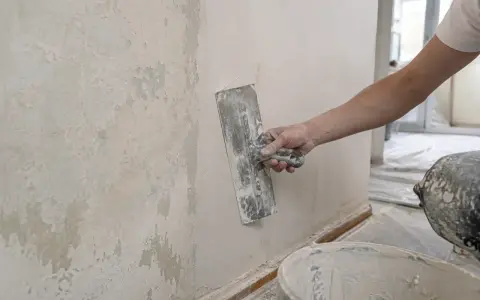 Ręka z kielnią nakłada gładź na chropowatą ścianę, pokazując, jak położyć gładź na ścianę dla idealnie równej powierzchni.