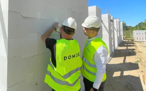Dwóch mężczyzn w kamizelkach odblaskowych i kaskach podczas inspekcji budowlanej. Jeden z nich sprawdza bloczki z betonu komórkowego.