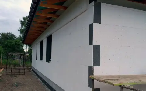 Budowa domu w toku. Ściany ocieplone grubym styropianem na placki, widoczne drewniane elementy dachu i okna.