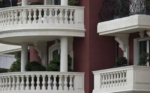 Eleganckie balustrady balkonowe z kamienia, z widocznym mocowaniem balustrady przez styropian, otaczają budynek o bordowej fasadzie.