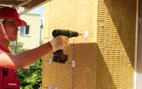 Pracownik w kasku montuje wełnę mineralną do ściany drewnianej za pomocą wkrętarki.