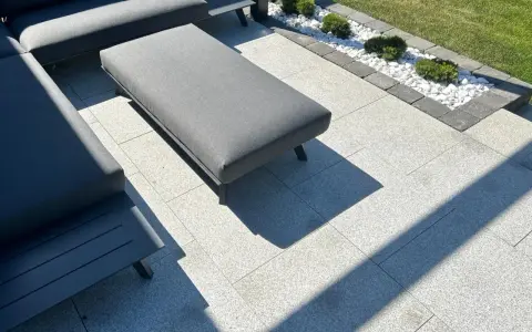 Nowoczesny taras z szarymi meblami, gdzie trwa układanie płyt tarasowych na podsypce. W tle rabata z białymi kamieniami i bukszpanami.