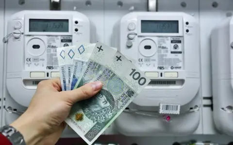 Dłoń trzyma polskie banknoty przed licznikiem energii elektrycznej. Brak licznika dwukierunkowego przy instalacji fotowoltaicznej może oznaczać brak rozliczenia nadwyżek.