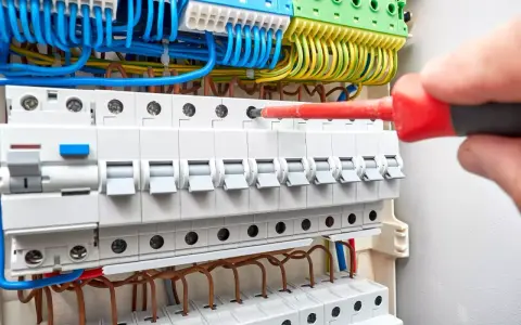 Ręka z czerwonym śrubokrętem dokręca śrubę w budowie rozdzielni elektrycznej. Widać bezpieczniki i kolorowe przewody.