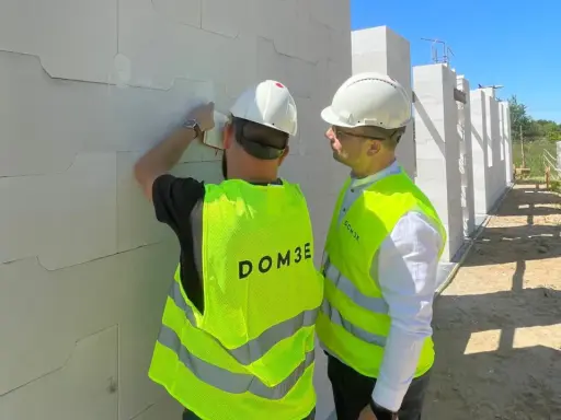 Dwóch mężczyzn w kamizelkach odblaskowych i kaskach podczas inspekcji budowlanej. Jeden z nich sprawdza bloczki z betonu komórkowego.