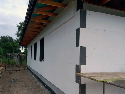 Budowa domu w toku. Ściany ocieplone grubym styropianem na placki, widoczne drewniane elementy dachu i okna.