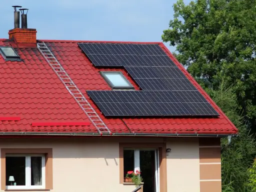 Panele fotowoltaiczne na czerwonym dachu domu. Drabina prowadzi do komina. Chcesz wiedzieć, jak sprawdzić ile prądu wyprodukowała fotowoltaika?