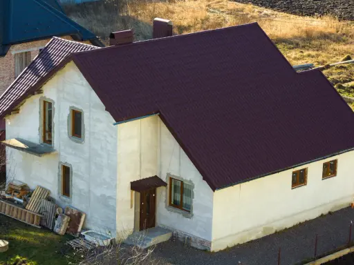 Nowy dom w zabudowie siedliskowej, z ciemnobordowym dachem i białymi ścianami, czeka na wykończenie.