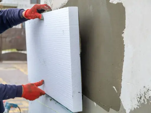 Ręce w czerwonych rękawicach przytwierdzają biały styropian do ściany. To dowód na świetne właściwości styropianu w izolacji termicznej.