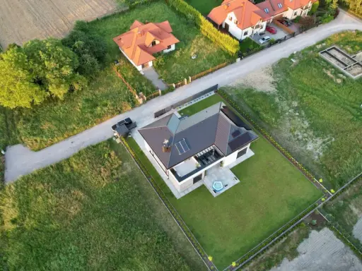 Widok z lotu ptaka na nowoczesny dom z panelami słonecznymi i basenem. Obok widać fundamenty nowego budynku, co sugeruje plan zagospodarowania przestrzennego dla konkretnej działki.
