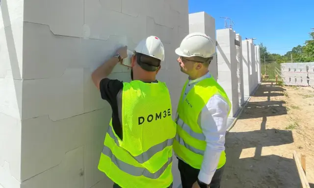 Dwóch mężczyzn w kamizelkach odblaskowych i kaskach podczas inspekcji budowlanej. Jeden z nich sprawdza bloczki z betonu komórkowego.