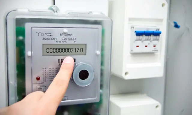 Palec wskazuje stan licznika energii elektrycznej: 00000000717.0 kWh. Obok widoczny wyłącznik.