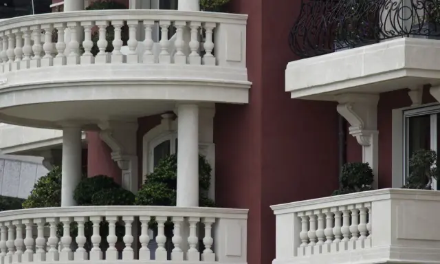 Eleganckie balustrady balkonowe z kamienia, z widocznym mocowaniem balustrady przez styropian, otaczają budynek o bordowej fasadzie.