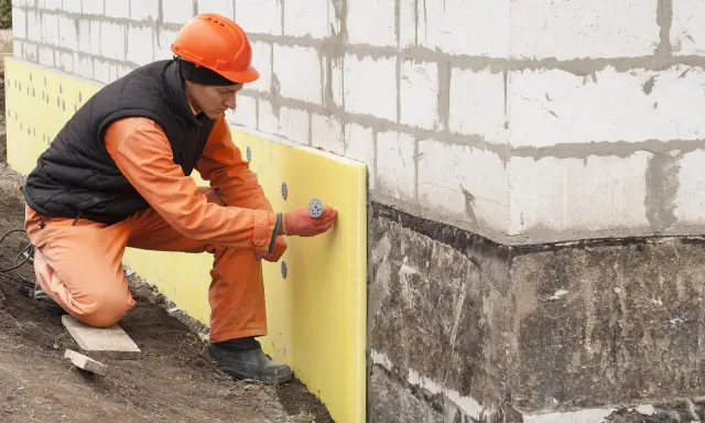 Budowlaniec mocuje żółty styropian do ściany fundamentowej. Czy styropian pije wodę? Nie, jest wodoodporny.