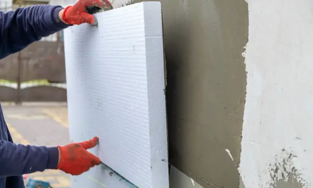 Ręce w czerwonych rękawicach przytwierdzają biały styropian do ściany. To dowód na świetne właściwości styropianu w izolacji termicznej.
