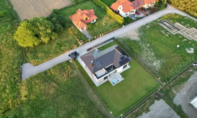 Widok z lotu ptaka na nowoczesny dom z panelami słonecznymi i basenem. Obok widać fundamenty nowego budynku, co sugeruje plan zagospodarowania przestrzennego dla konkretnej działki.