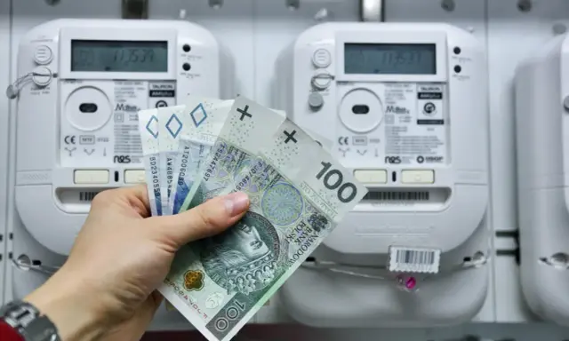Dłoń trzyma polskie banknoty przed licznikiem energii elektrycznej. Brak licznika dwukierunkowego przy instalacji fotowoltaicznej może oznaczać brak rozliczenia nadwyżek.