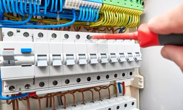 Ręka z czerwonym śrubokrętem dokręca śrubę w budowie rozdzielni elektrycznej. Widać bezpieczniki i kolorowe przewody.