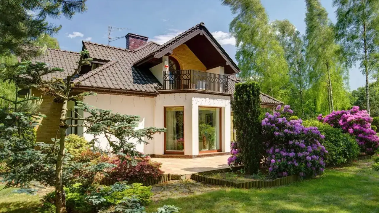 Dom z ryzalitem, balkonem i kwitnącymi rododendronami w ogrodzie.