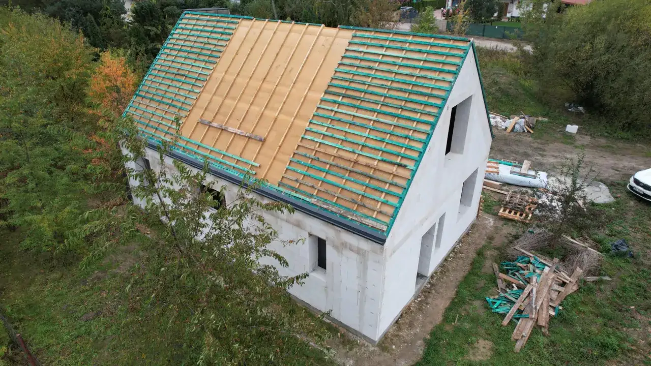 Rozbudowa domu w toku. Białe ściany z bloczk&oacute;w, dach w trakcie prac, widoczne drewniane łaty i zielone membrany.