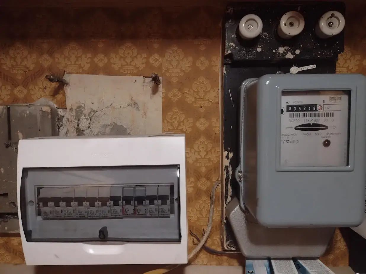 Budowa rozdzielni elektrycznej: licznik energii i biała obudowa z bezpiecznikami na tle tapety.