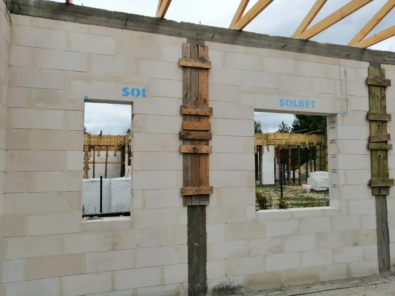 Ściana z bloczk&oacute;w z dwoma otworami okiennymi. Nad jednym z nich widoczna jest betonowa belka nad oknem, podparta drewnianymi stemplami.